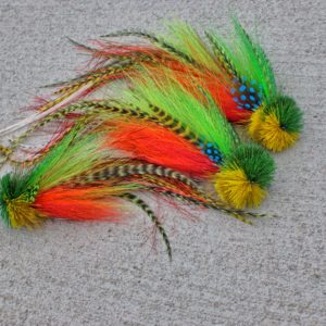 A collection of brightly colored fly fishing lures by Urban Fly Co., featuring combinations of green, yellow, and orange feathers with striped and spotted accents, displayed on a concrete surface.