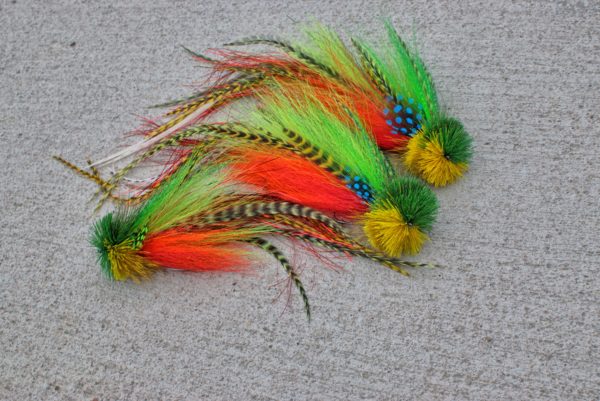 A collection of brightly colored fly fishing lures by Urban Fly Co., featuring combinations of green, yellow, and orange feathers with striped and spotted accents, displayed on a concrete surface.