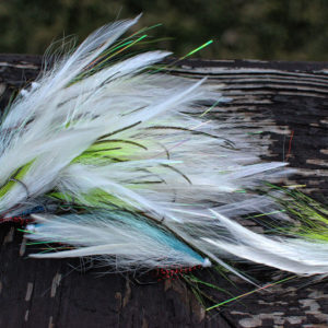 A collection of musky fly fishing lures by Urban Fly Co., named 'Lefty Feature,' featuring a combination of white, green, and blue feathers, displayed on a weathered wooden surface with a blurred natural background.