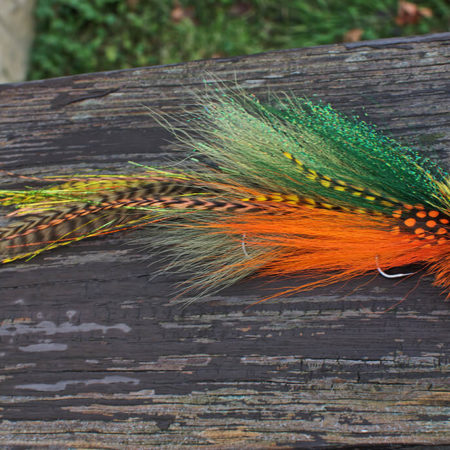 A vibrant fly fishing lure by Urban Fly Co., named 'Perch Double,' featuring a combination of green, orange, and yellow feathers with striped and spotted accents, and a bushy yellow and orange head, displayed on a weathered wooden surface with a blurred natural background.