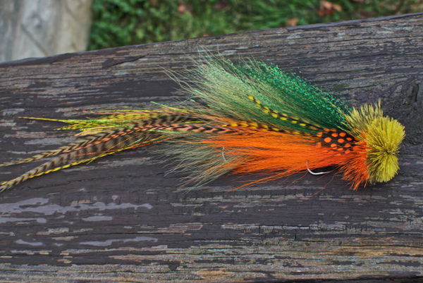 A vibrant fly fishing lure by Urban Fly Co., named 'Perch Double,' featuring a combination of green, orange, and yellow feathers with striped and spotted accents, and a bushy yellow and orange head, displayed on a weathered wooden surface with a blurred natural background.