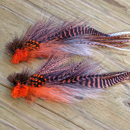 A pair of detailed fly fishing lures by Urban Fly Co., featuring a combination of orange, black, and brown feathers with striped and spotted accents, and bushy orange and black heads, displayed on a wooden surface.