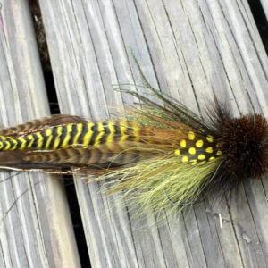 A detailed fly fishing lure by Urban Fly Co., featuring a combination of brown, yellow, and green feathers with yellow polka dot accents and a bushy brown head, displayed on a wooden surface.