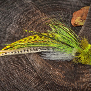 A detailed fly fishing lure by Urban Fly Co., featuring a combination of yellow, green, and white feathers with striped accents, and a bushy yellow head, displayed on a tree stump with a natural background.