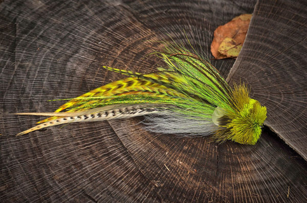 A detailed fly fishing lure by Urban Fly Co., featuring a combination of yellow, green, and white feathers with striped accents, and a bushy yellow head, displayed on a tree stump with a natural background.