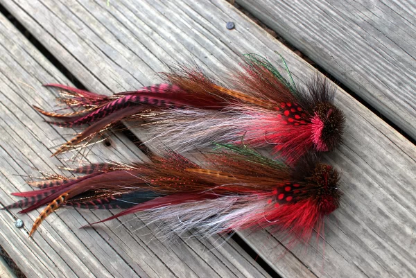 Pair of Urban Fly Co. lures with red and brown feathers, designed for fly fishing, arranged on a wooden surface.