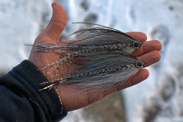 Urban Fly Co. Shad Bendback fishing lures, featuring a detailed feather design with black, white, and reflective elements, held in a hand against a snowy background.