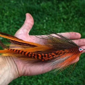 Urban Fly Co. Sucker Single Minnow fishing lure with vibrant orange and black feathers, displayed on a hand with a green grass background.