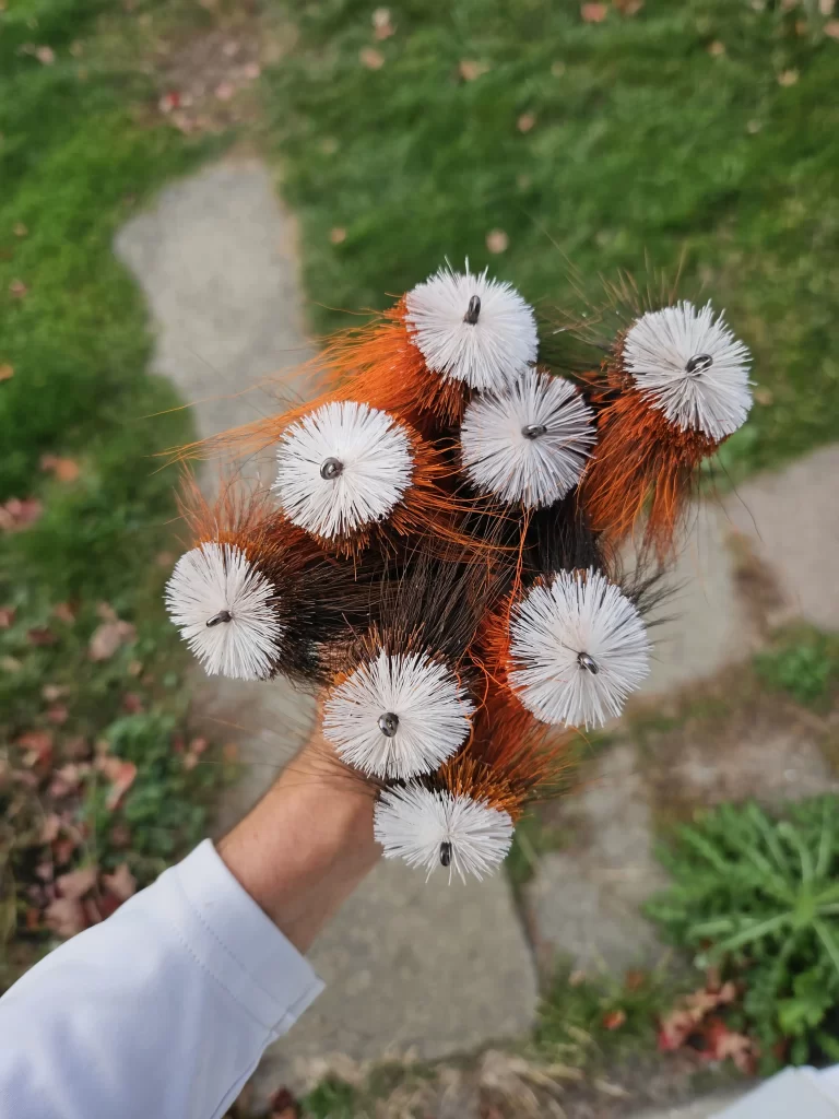 Urban Fly Co. cluster of WF Sucker fishing lures, showcasing detailed craftsmanship with orange, brown, and white feather patterns, held above a green lawn background.