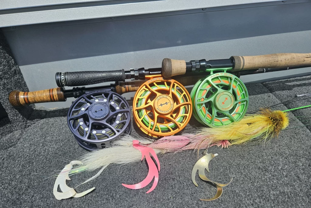 Iconic Fly fishing gear from Urban Fly Co. featuring three fly reels in different colors (black, orange, and green), fly rods, and a variety of lures laid out on a carpeted surface.