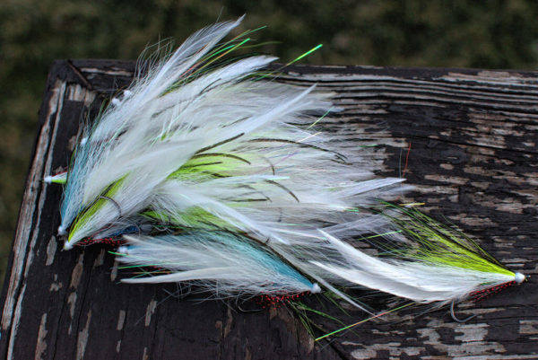 A collection of musky fly fishing lures by Urban Fly Co., named 'Lefty Feature,' featuring a combination of white, green, and blue feathers, displayed on a weathered wooden surface with a blurred natural background.