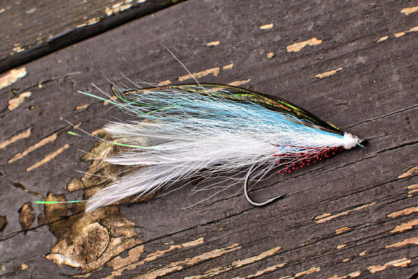 A single fly fishing lure by Urban Fly Co., named 'Lefty' featuring a combination of white, blue, and brown feathers, displayed on a weathered wooden surface with a blurred natural background.