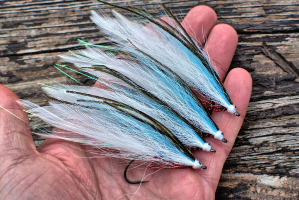 A collection of fly fishing lures by Urban Fly Co., named 'Lefty' featuring a combination of white, green, and blue feathers, displayed in a person's hand on a weathered wooden surface.