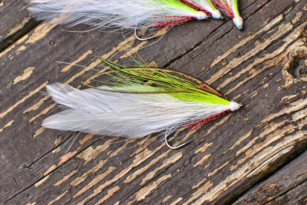 A single fly fishing lure by Urban Fly Co., named 'Lefty,' featuring a combination of white, green, and brown feathers, displayed on a weathered wooden surface with additional lures partially visible at the top