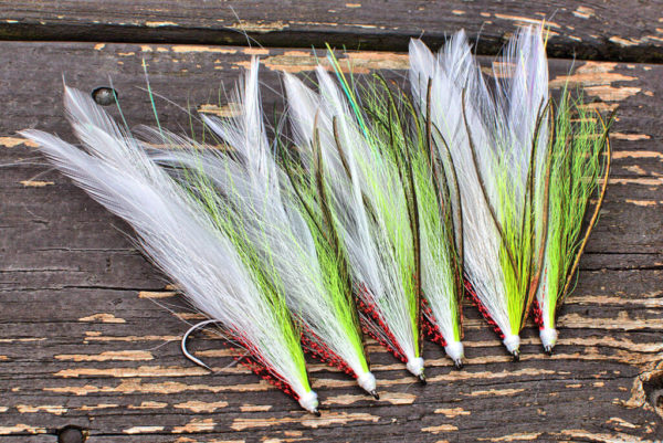 A collection of musky fly fishing lures by Urban Fly Co., named 'Lefty Two,' featuring a combination of white, green, and brown feathers, neatly arranged on a weathered wooden surface.