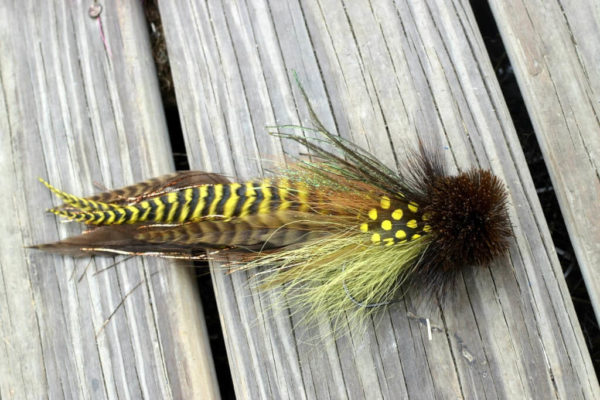 A detailed fly fishing lure by Urban Fly Co., featuring a combination of brown, yellow, and green feathers with yellow polka dot accents and a bushy brown head, displayed on a wooden surface.