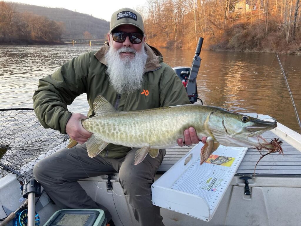 Angler holding a large muskie caught using an Urban Fly Co. lure, seated on a boat with a scenic river background.