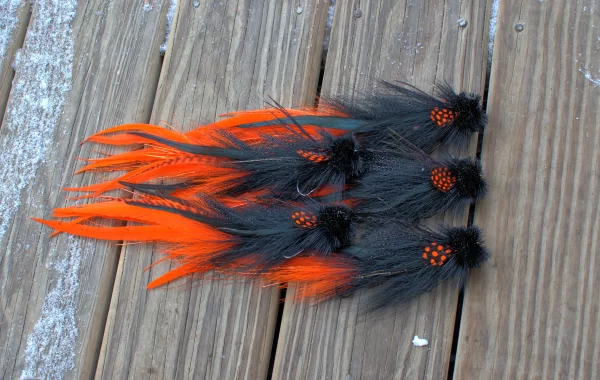 Set of Urban Fly Co. lures with black and vibrant orange feathers, designed for fly fishing, arranged on a wooden surface with a dusting of snow.