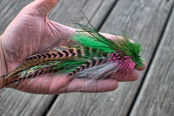 Hand holding a vibrant green and pink Urban Fly Co. lure with striped feathers and a bushy head, showcasing its detailed craftsmanship.