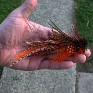 Hand holding an orange Urban Fly Co. lure with detailed feathers and a bushy head, showcasing its intricate design.