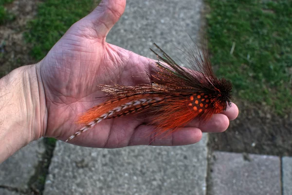 Hand holding an orange Urban Fly Co. lure with detailed feathers and a bushy head, showcasing its intricate design.