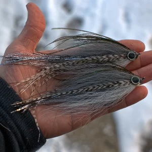 Urban Fly Co. Shad Bendback fishing lures, featuring a detailed feather design with black, white, and reflective elements, held in a hand against a snowy background.