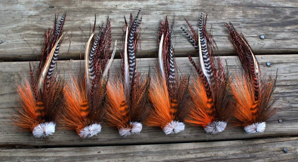 Urban Fly Co. collection of orange and brown single Buford fishing lures displayed on a wooden surface, showcasing detailed feather patterns and textures.