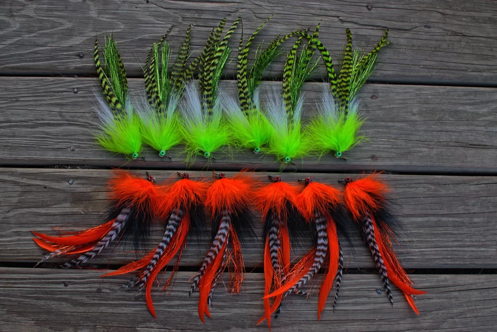 Two rows of Urban Fly Co. fly fishing lures arranged on a wooden surface; the top row features lures with green and yellow feathers, while the bottom row features lures with black and orange feathers.