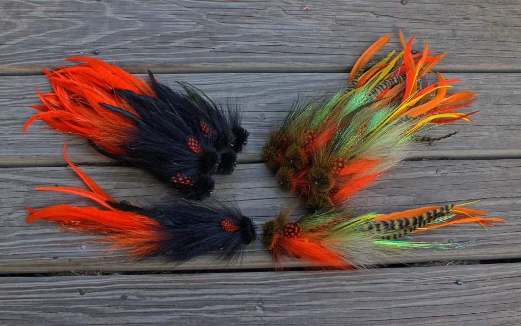 Four groups of Urban Fly Co. fly fishing lures arranged on a wooden surface, featuring feathers in shades of black, orange, green, and brown.